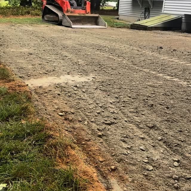 Gravel driveway being graded by a small orange skid steer, with green grass to the left and a building in the background.