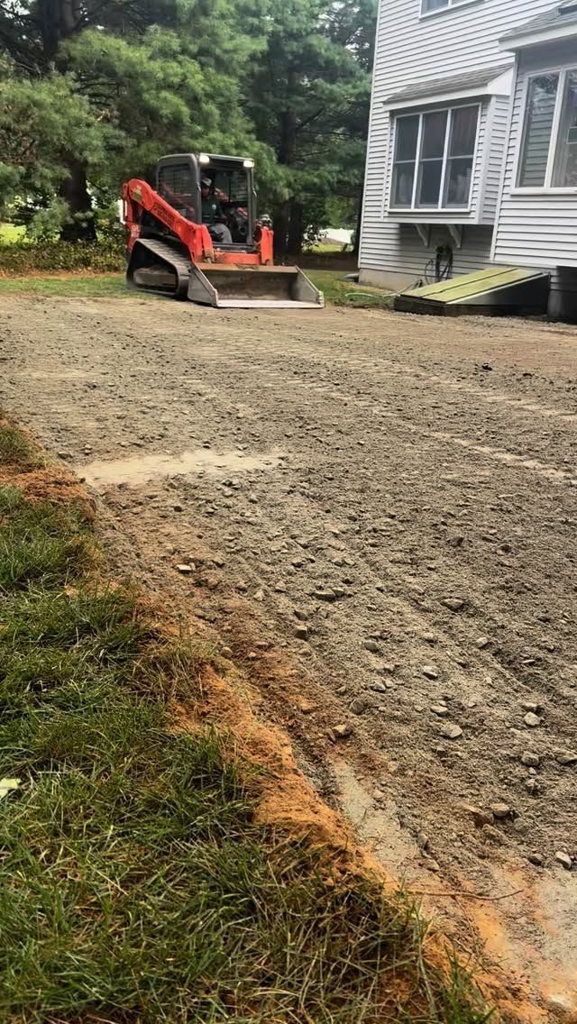 Driveway under construction with orange tractor near a house. Ground is uneven, with dirt and gravel.
