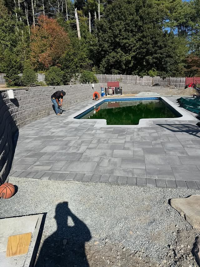 Poolside paving: a person works near a rectangular pool filled with green water. Gray pavers and gravel surround.