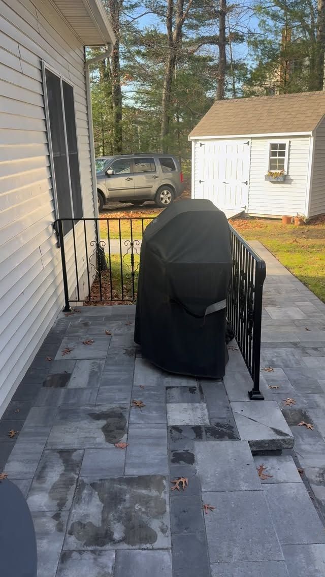 Patio with black covered grill, black railing, white shed and car.