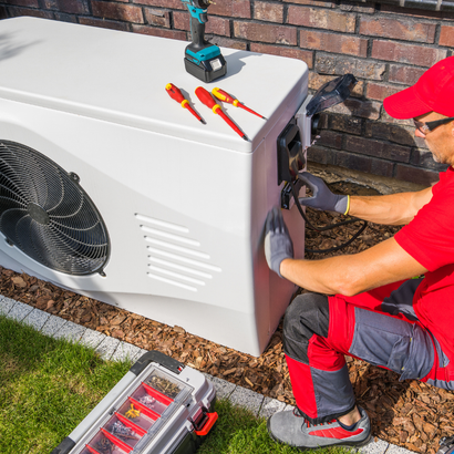 A person in red overalls installs a heat pump outdoors, using tools on the unit, next to a brick wall.