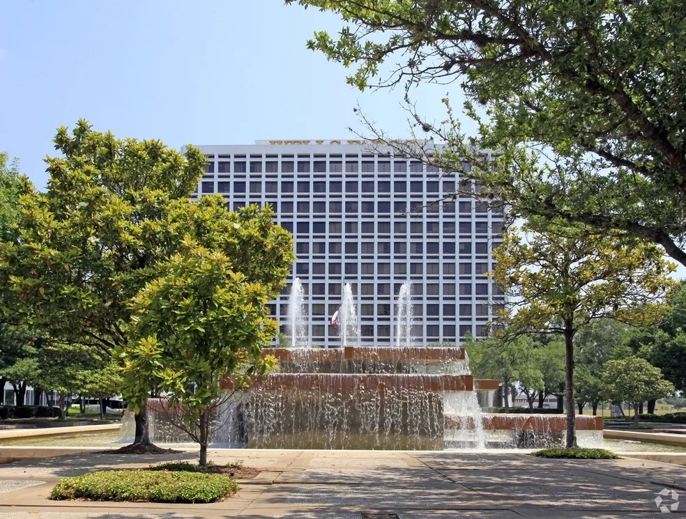 Fountain with tiered water features in front of a grid-patterned office building, framed by trees under a blue sky.