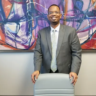 Man in suit stands, hands on a chair, in front of abstract painting.