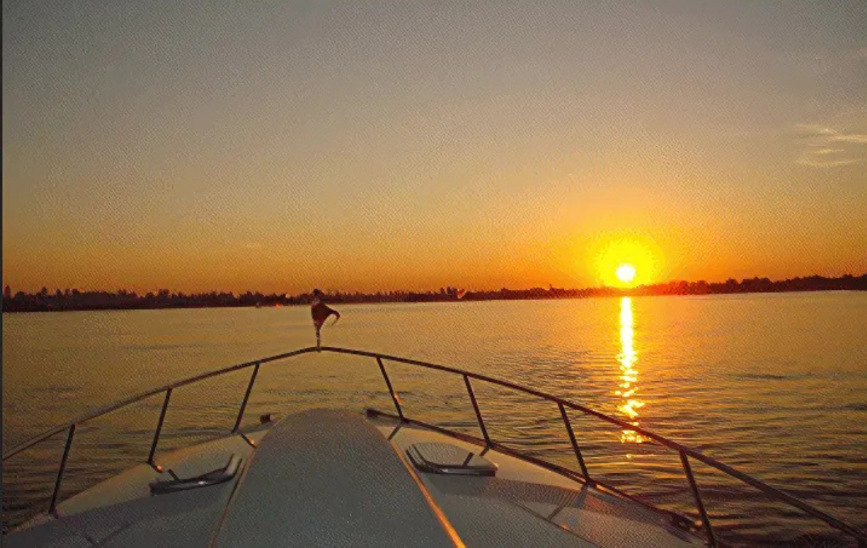 A boat travels on calm water toward a sunset. The sky is orange and yellow, reflecting on the water.