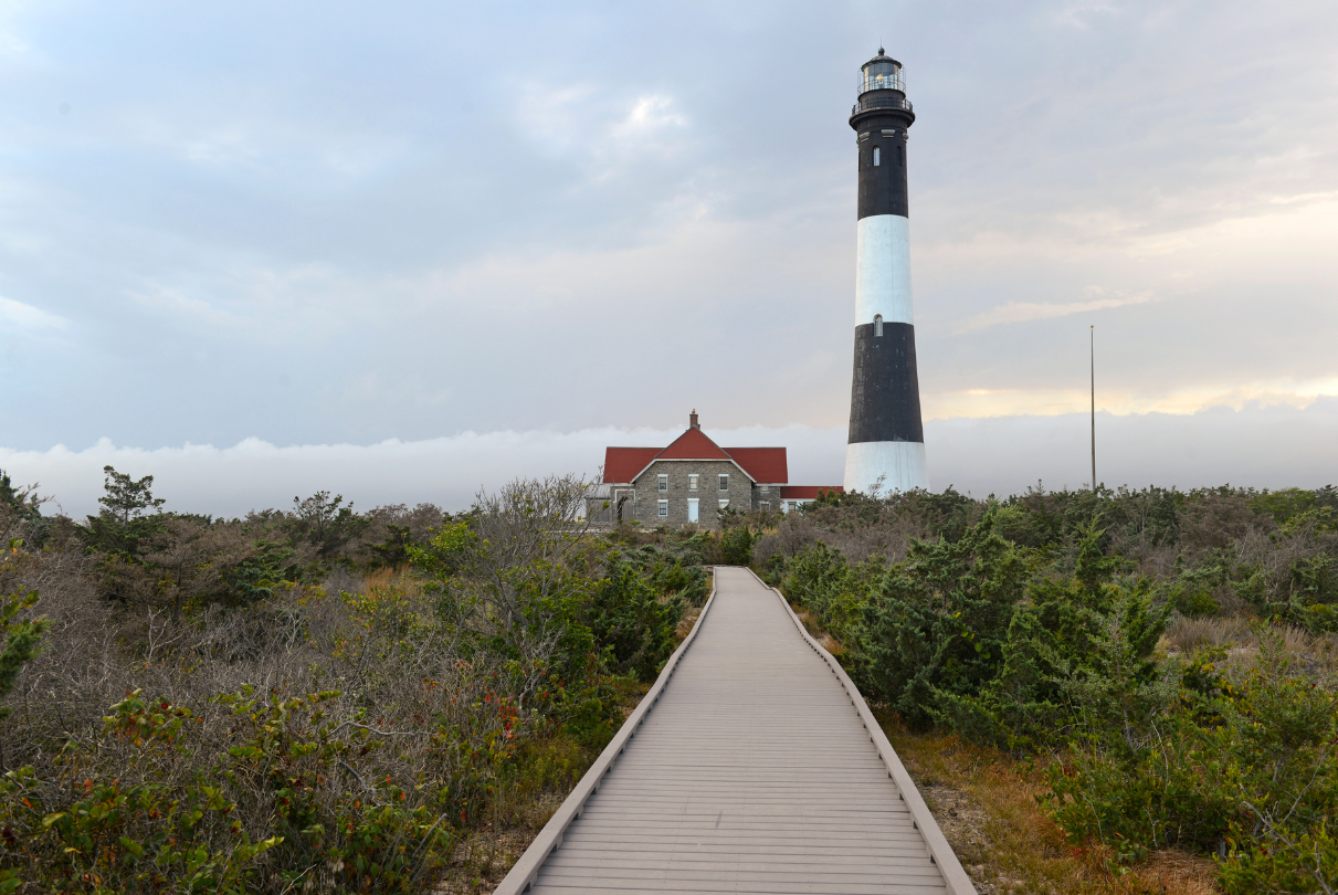 Wooden boardwalk leading to a tall black and white lighthouse and a red-roofed building on a cloudy day.
