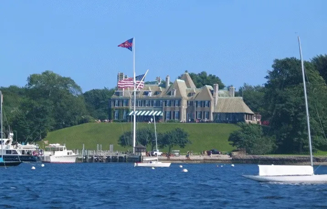 Large stone mansion on a grassy hill overlooking a harbor. Boats and a flagpole with flags are in the foreground, clear blue sky.