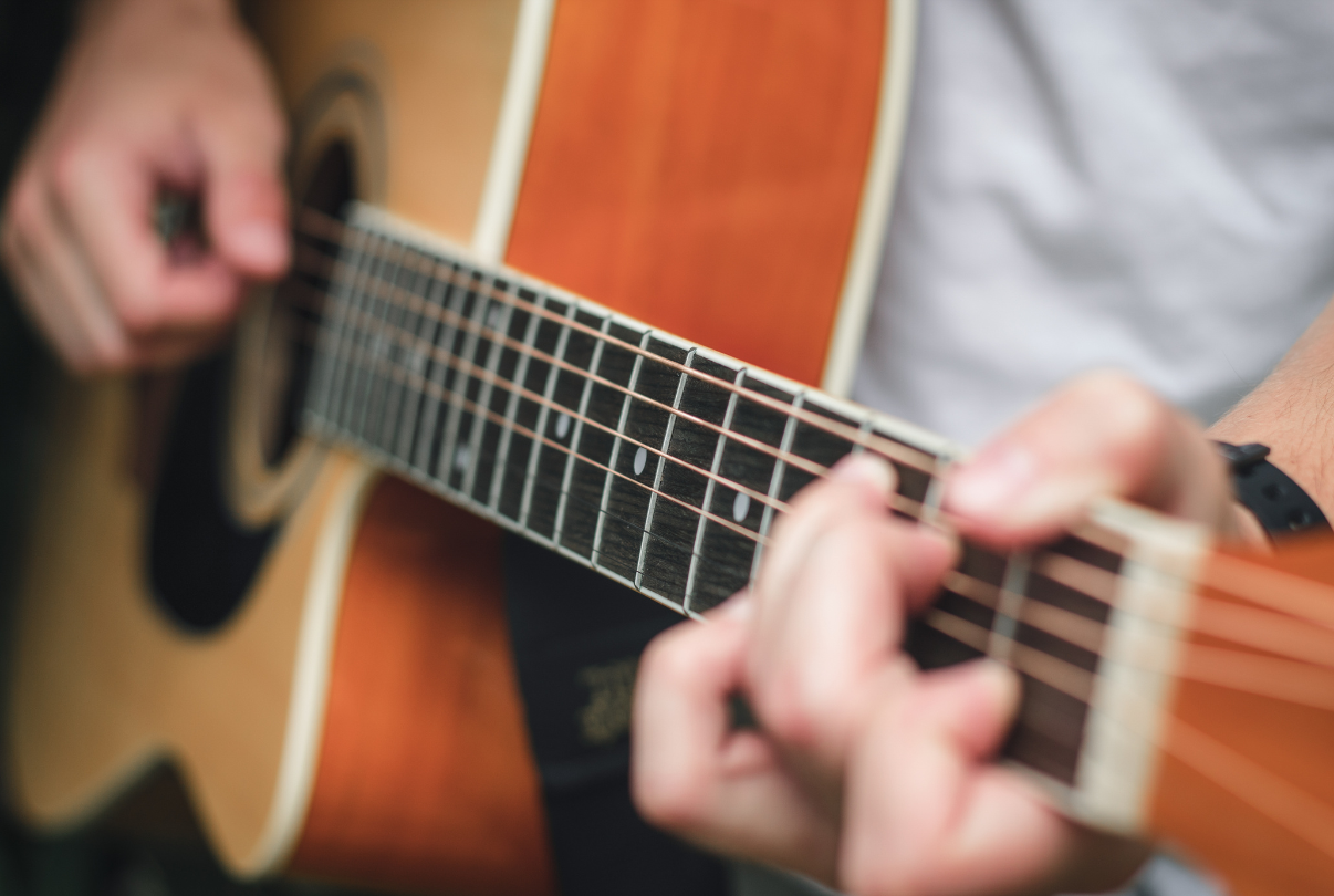 Person playing an acoustic guitar, hand strumming the strings. Blurred background of a stage setup.