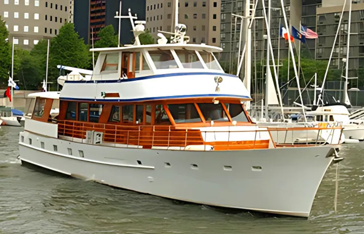White and wooden motor yacht on the water; the boat is moving, with buildings and other boats visible in the background.