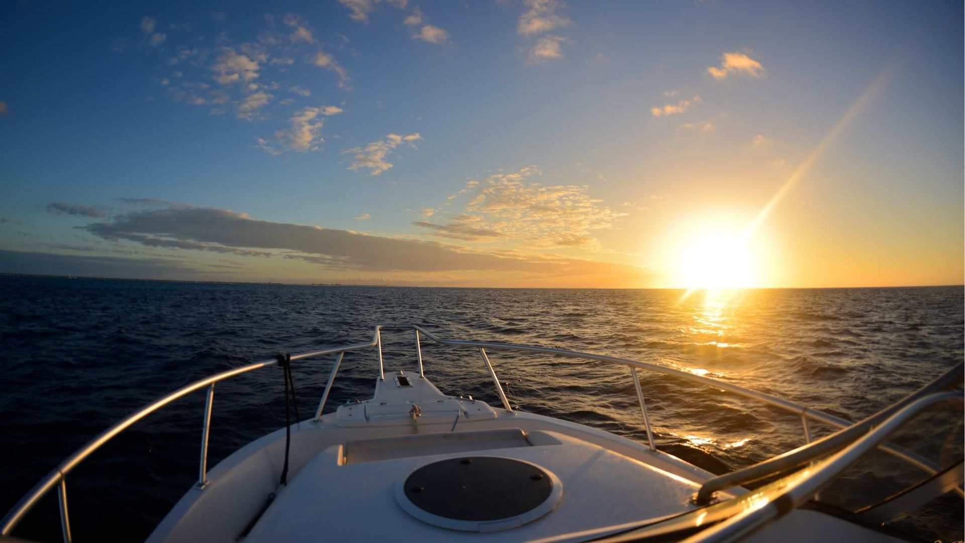 A boat sailing towards a bright sunset over the ocean. The sky is a mix of blue and gold.