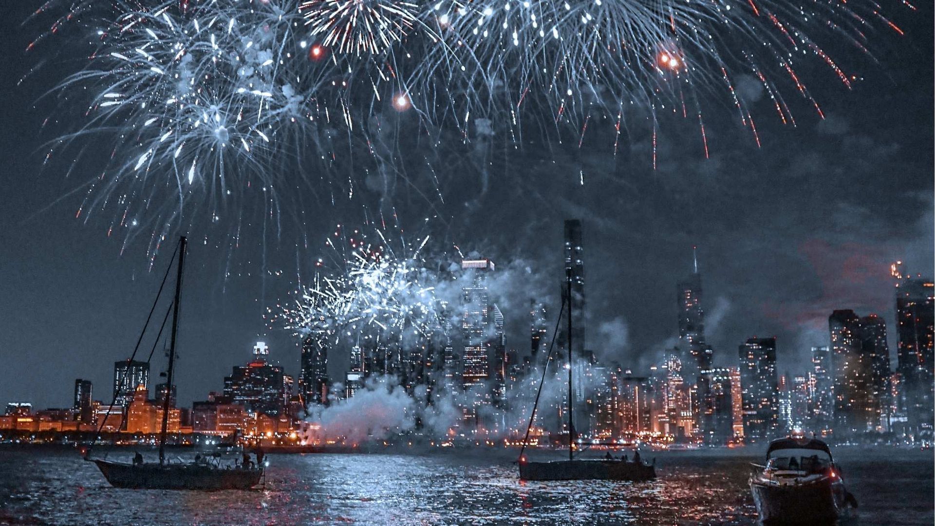 Fireworks explode over a city skyline at night, reflecting in the water with boats in the foreground.