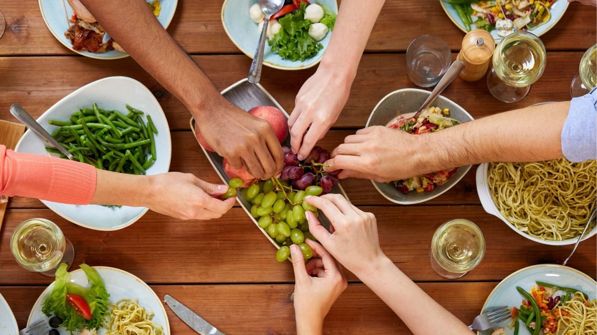 Hands reaching for a bowl of grapes at a table set with various dishes and drinks, a dining scene.
