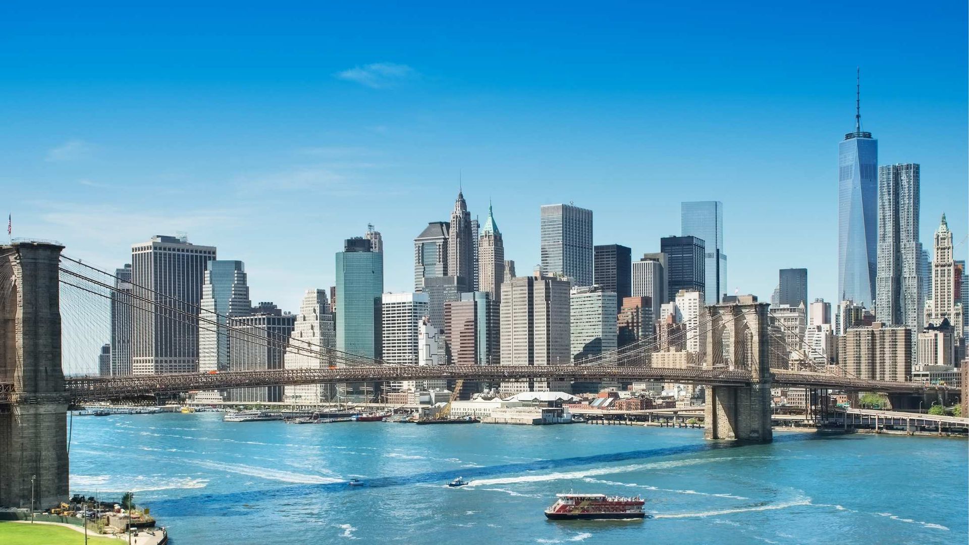 New York City skyline with the Brooklyn Bridge in the foreground, under a clear blue sky. Skyscrapers and water visible.
