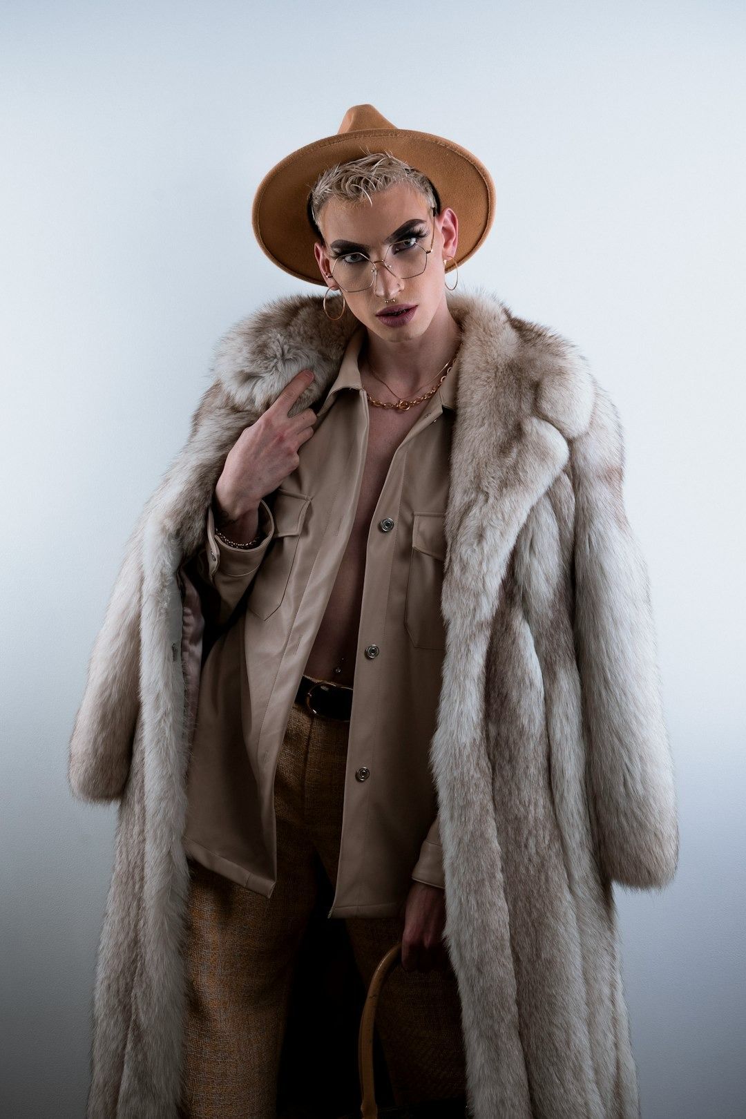 Person in tan shirt, fur coat, hat, and glasses, holding a basket, against a white wall.