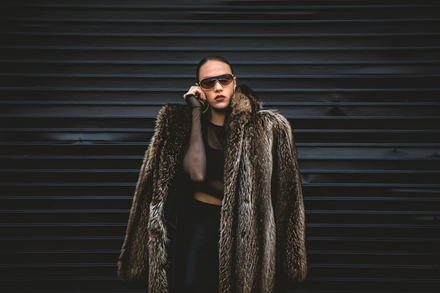 A woman wearing a fur coat and sunglasses is standing in front of a black wall.