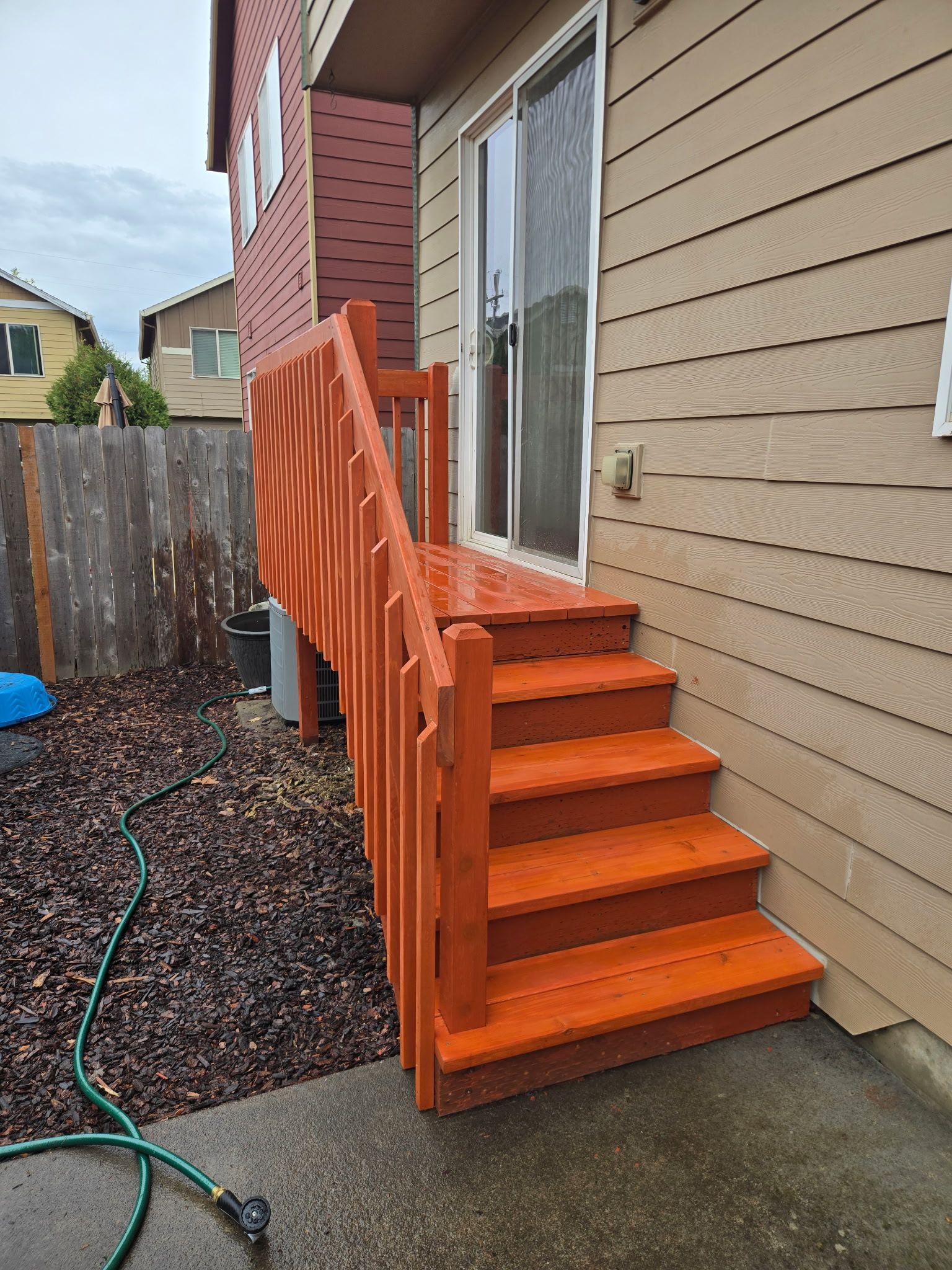 Orange wooden stairs and railing beside a beige house, leading to a back door in a narrow side yard.