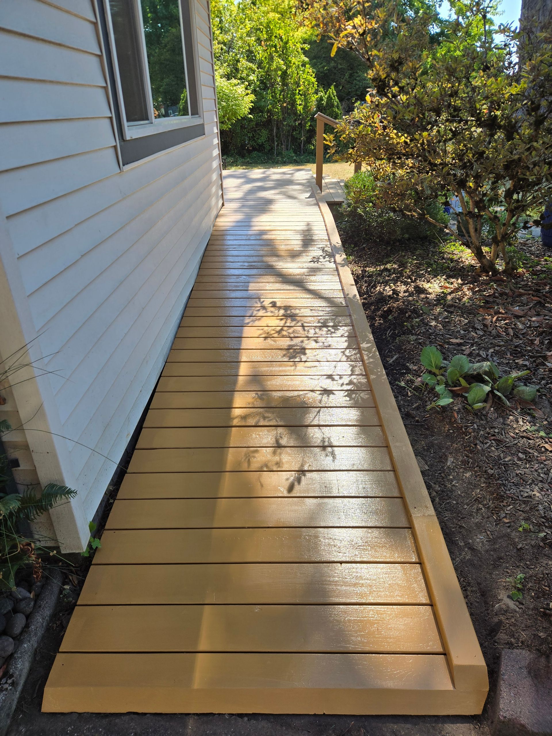 Wooden walkway beside a white house, bordered by mulch and shrubs, leading toward a sunny yard