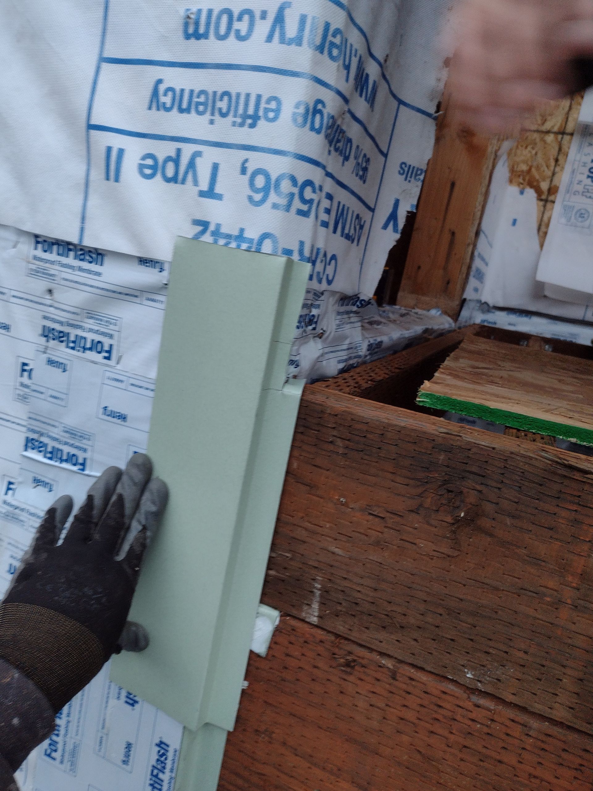 Gloved hand holding a white trim board beside a wood-framed wall under house wrap