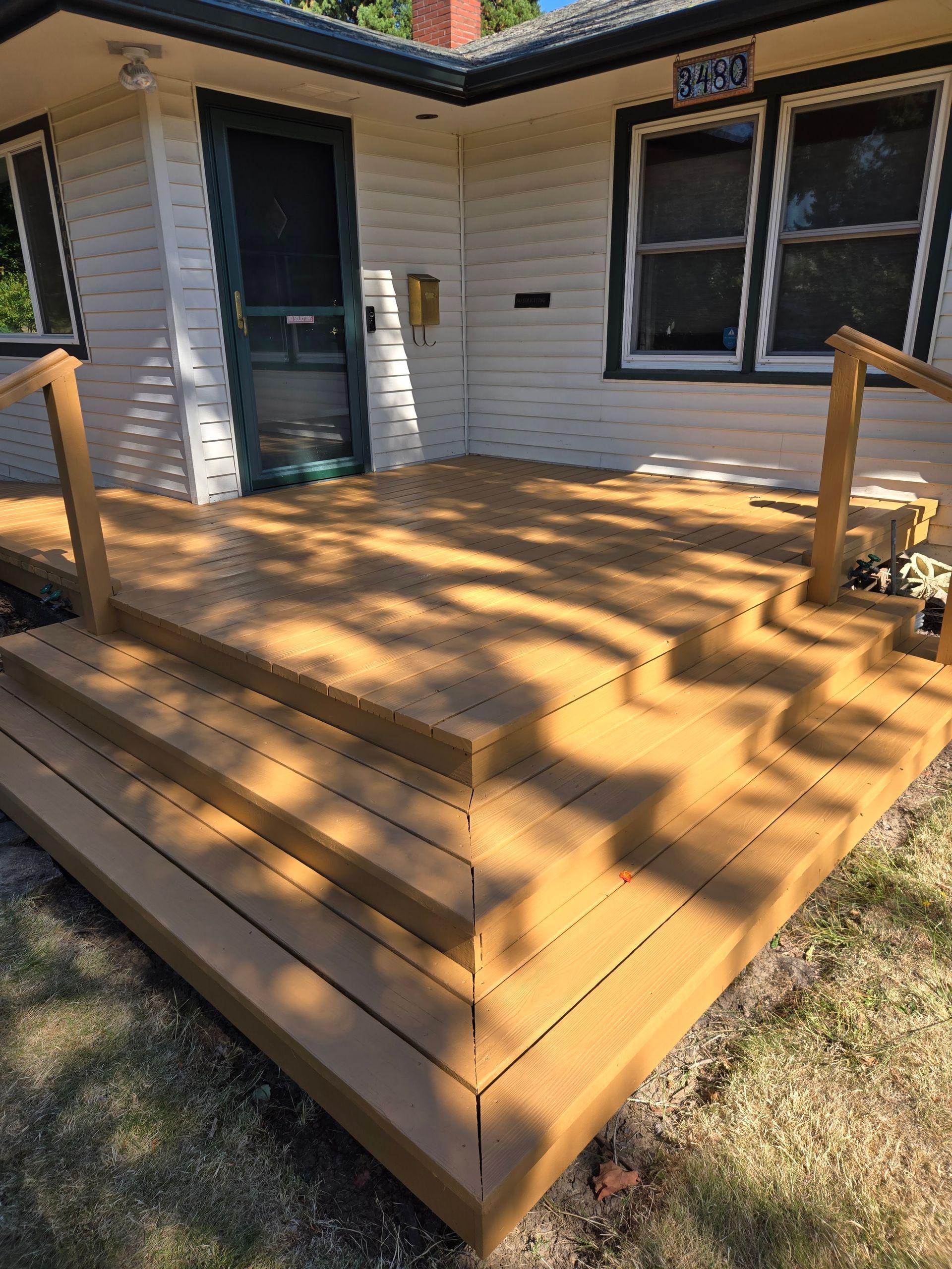 Wooden front porch and steps attached to a white house, with a door and window in sunlight.