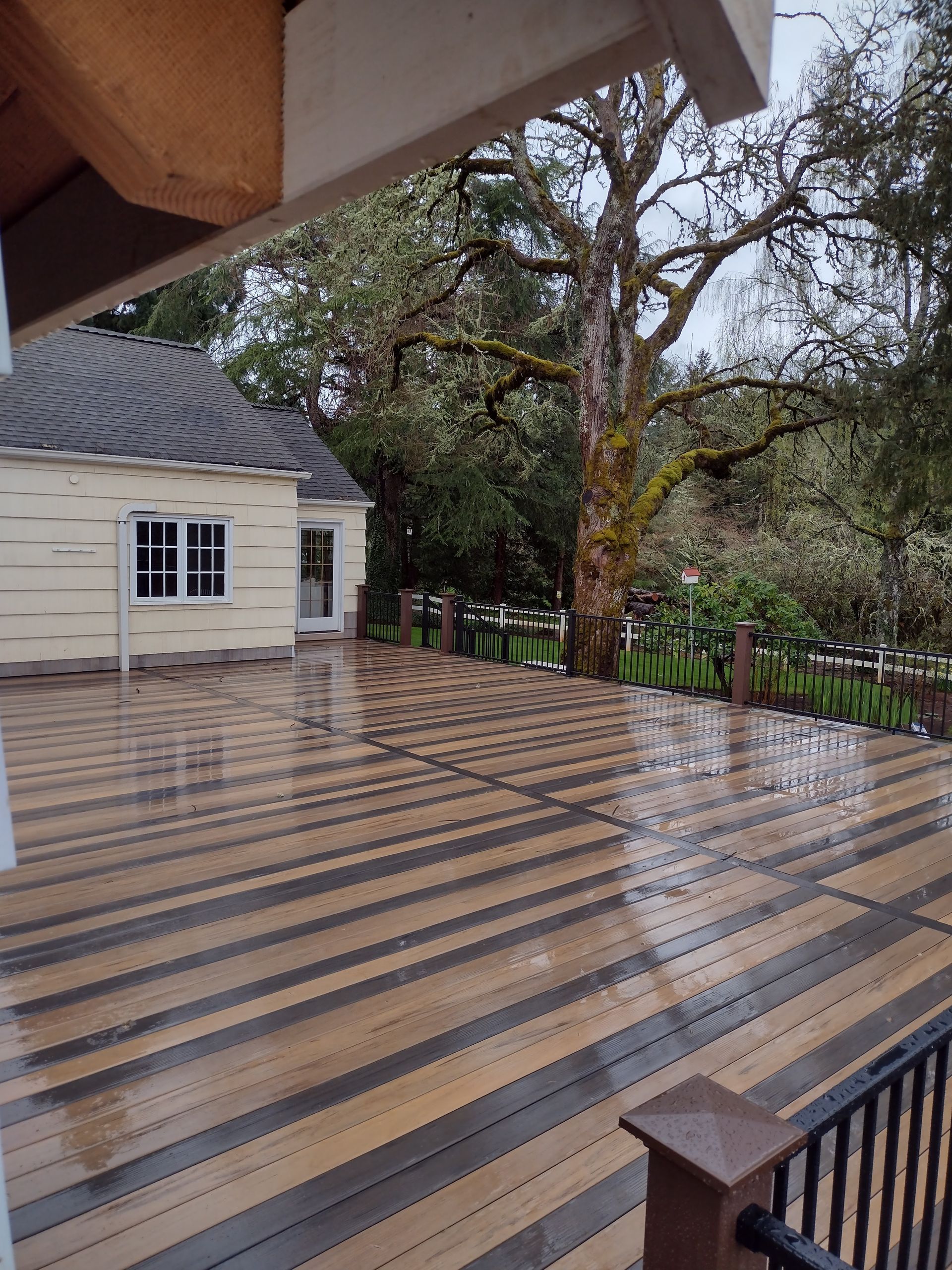 Rain-soaked striped wooden deck beside a white building and black railing, with trees beyond.