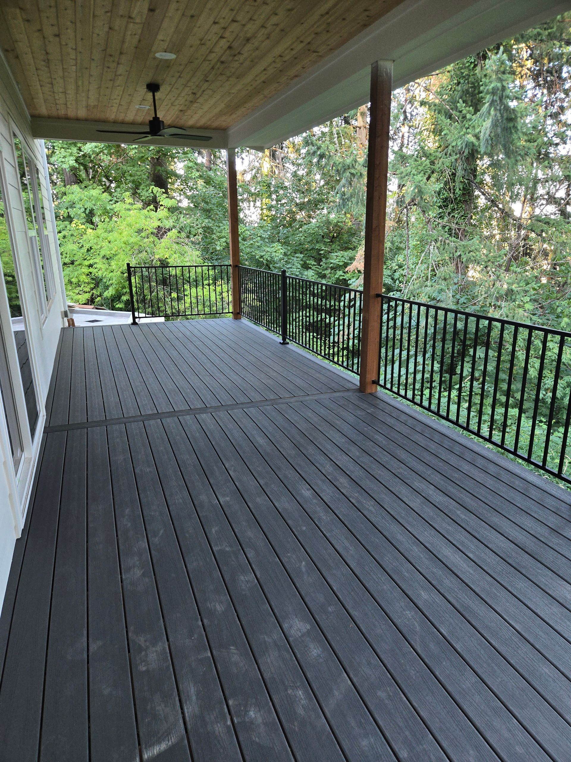 Covered wooden porch with gray floorboards, railing, and a ceiling fan overlooking green trees