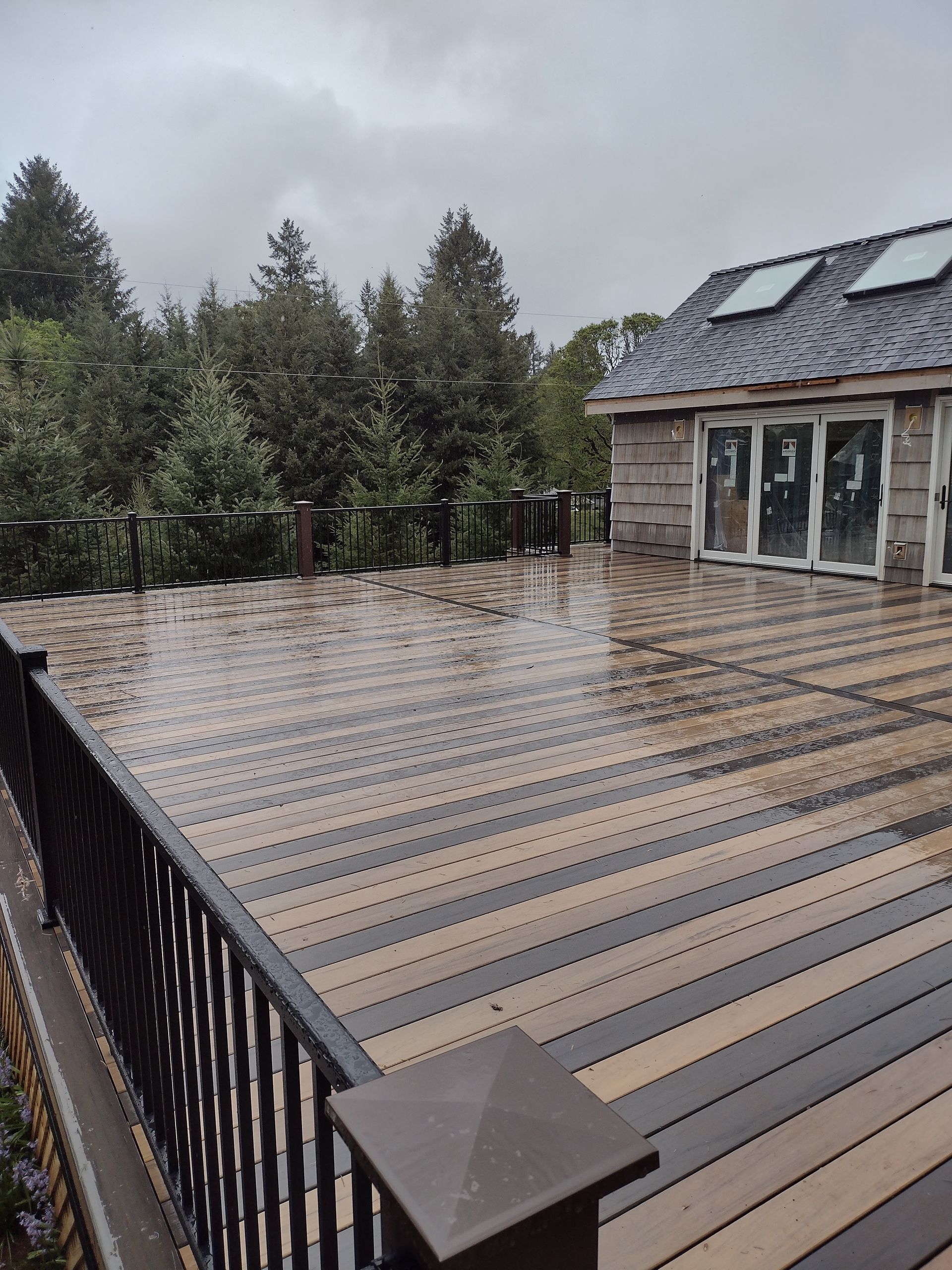 Snowy deck with railing beside a house and evergreen trees under a cloudy sky