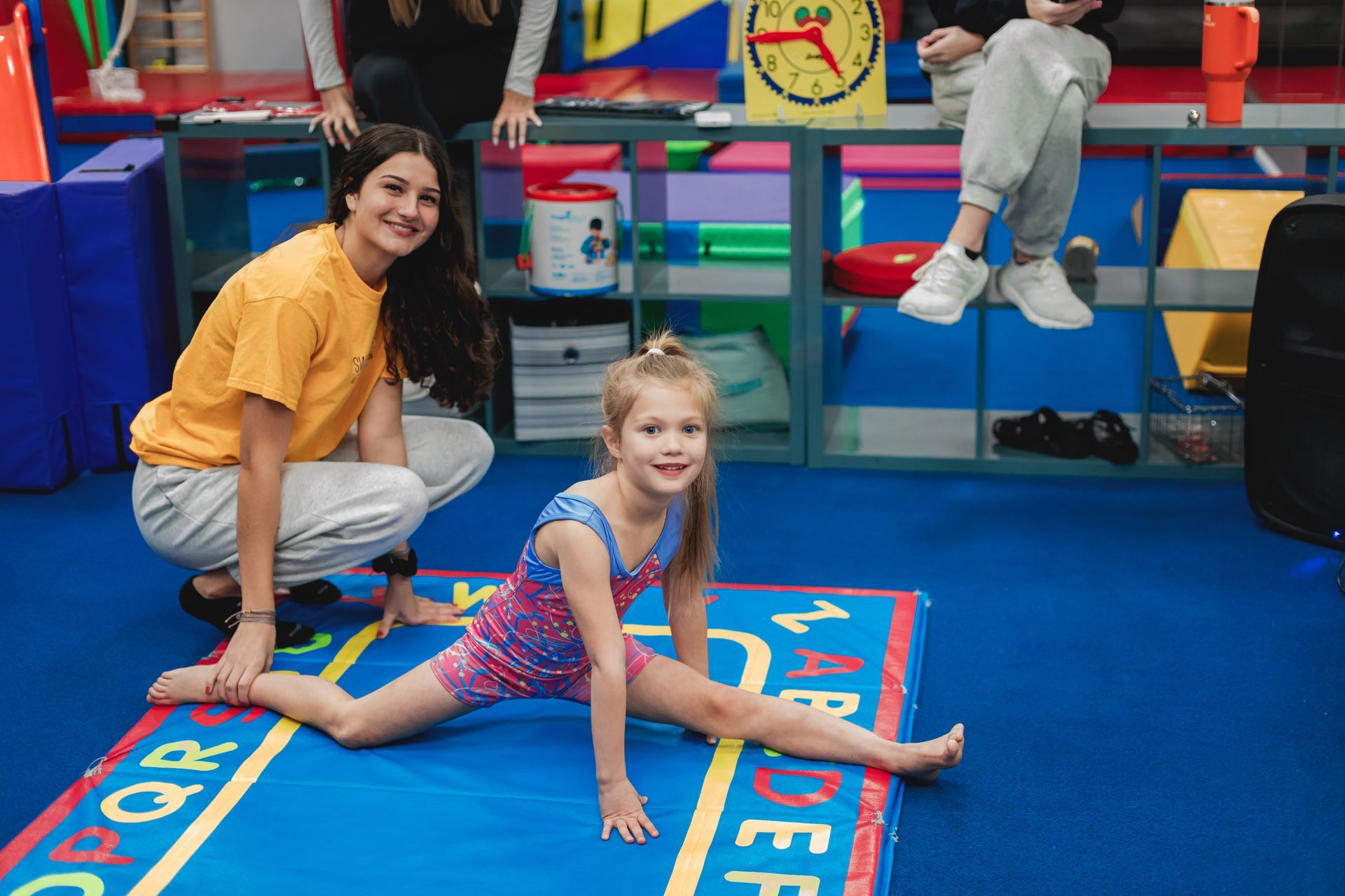 A woman is kneeling next to a little girl who is doing splits on a mat.