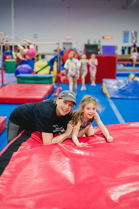A woman and a little girl are playing on a red mat in a gym.