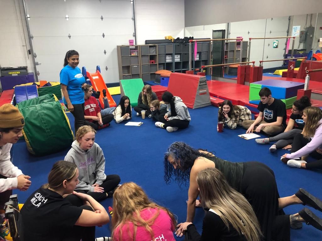 A group of people are sitting in a circle on the floor in a gym.