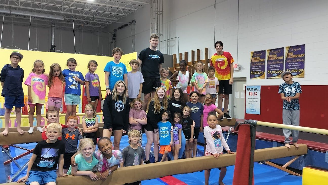 A group of children are standing on a balance beam in a gym.