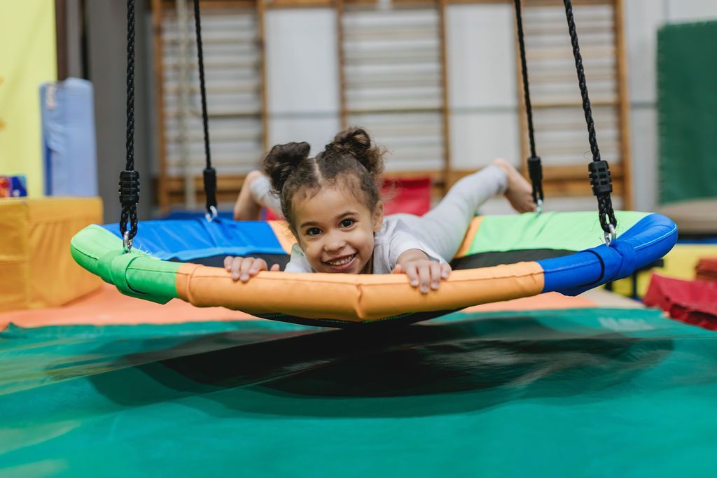 A little girl is laying on a colorful swing in a gym.