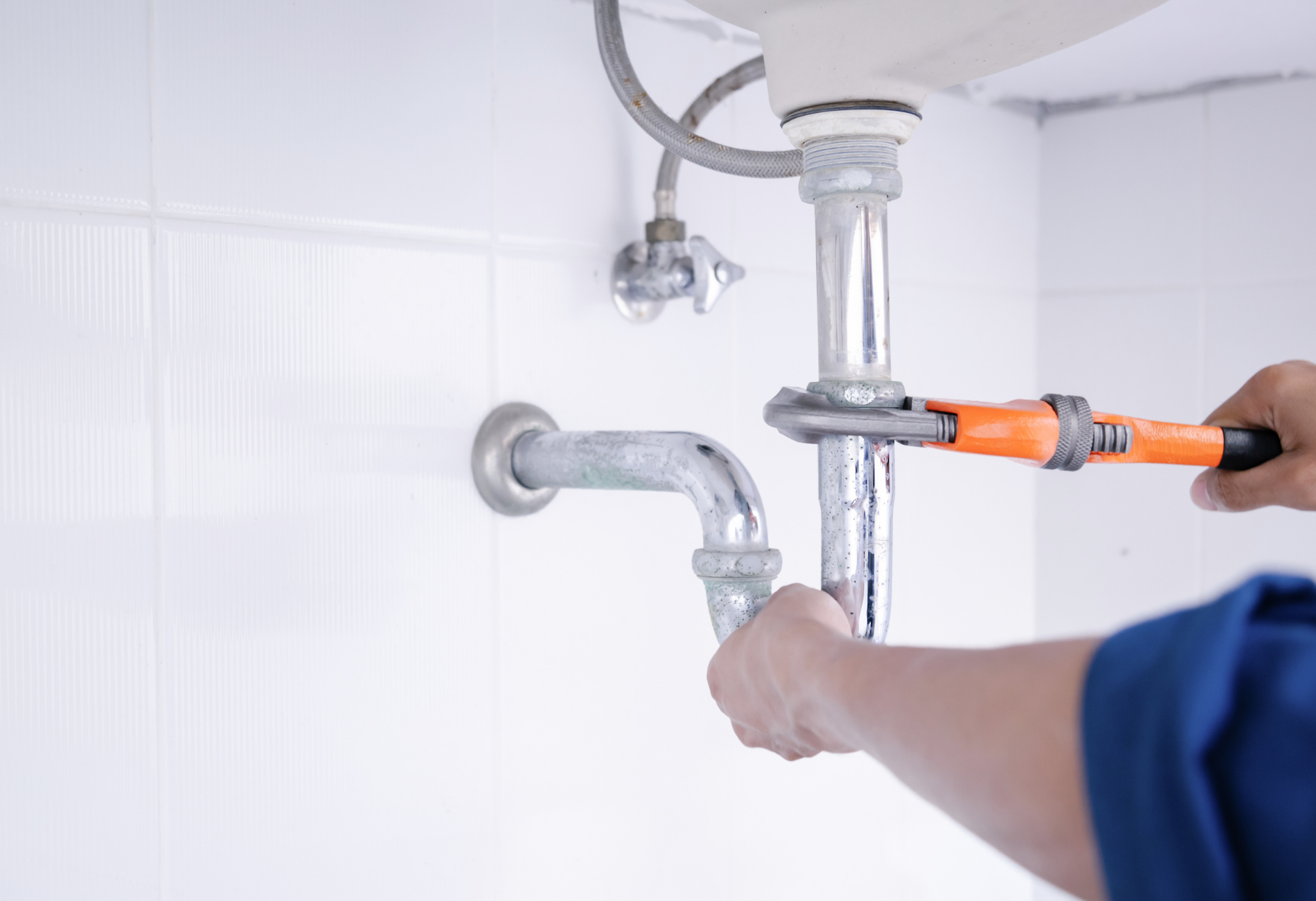 Plumber using a wrench to work on a chrome pipe under a white sink.