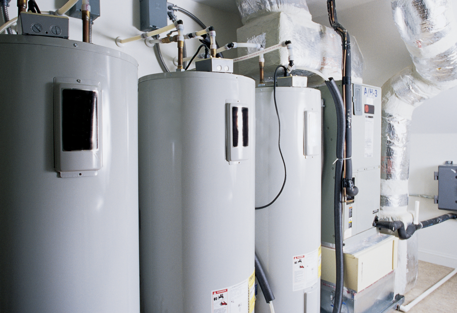 Three water heaters in a utility room. Gray tanks with electrical components, ductwork, and pipes.