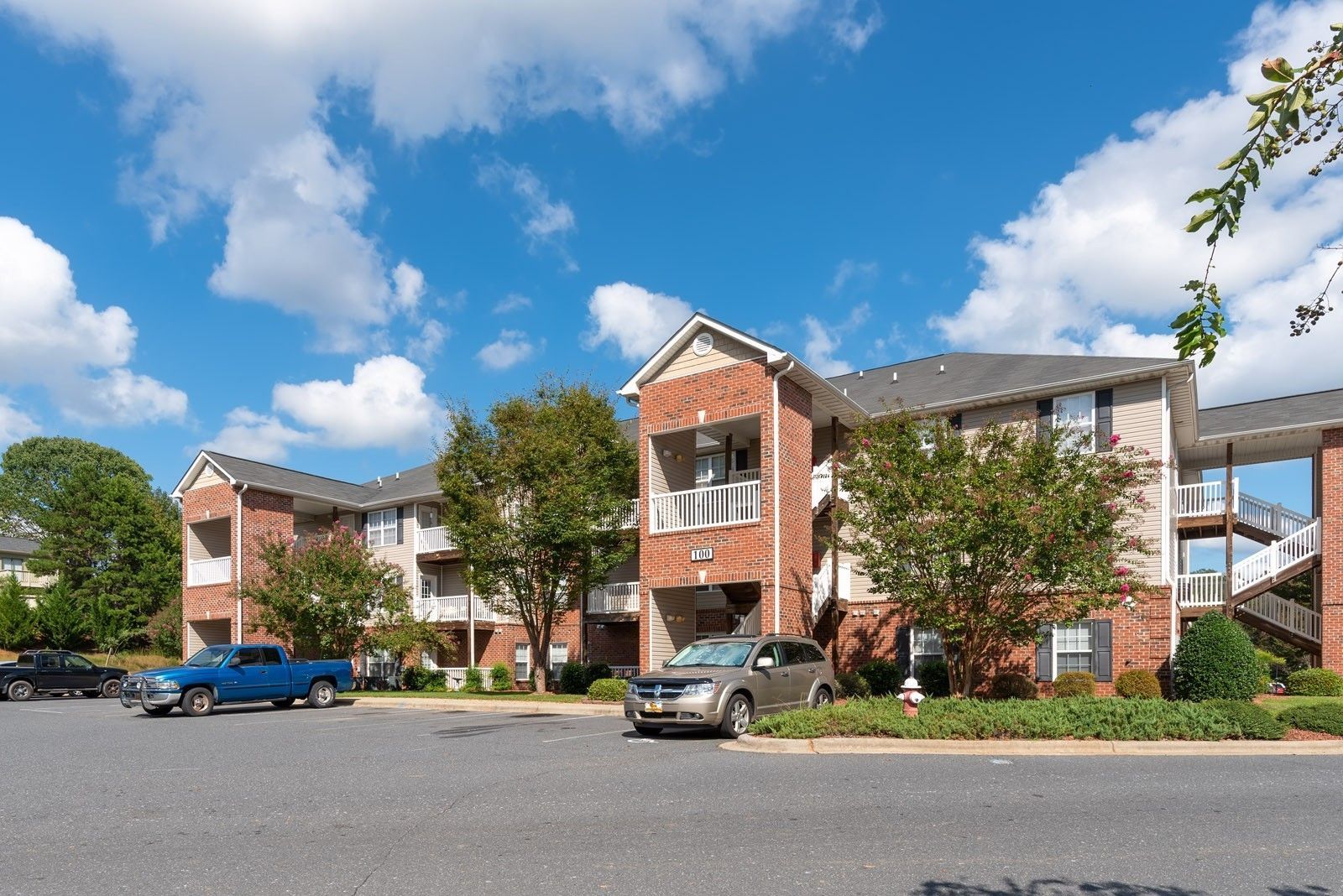 Apartment building with brick accents, tan siding, and balconies under a blue sky with clouds. Cars are parked out front.