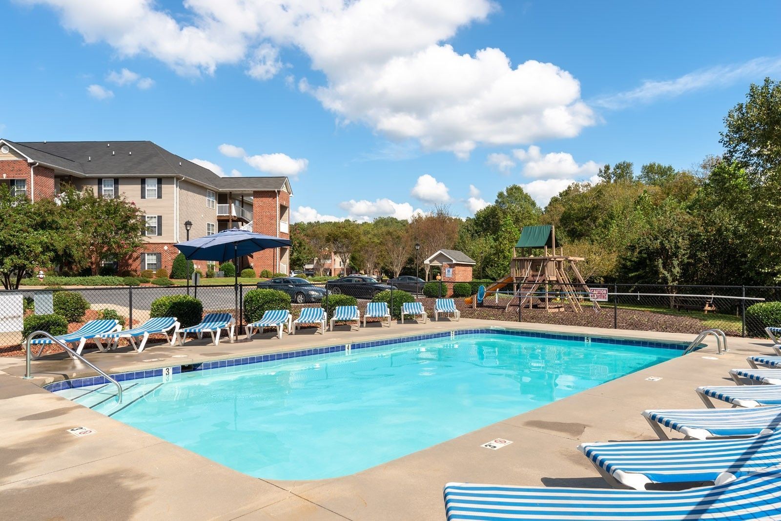 Swimming pool surrounded by lounge chairs, playground, and multi-story apartment building under a blue sky.