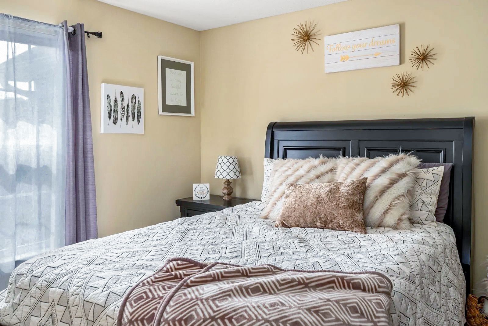 Bedroom with bed, nightstand, art, and window with sheer curtain. Light tan walls.
