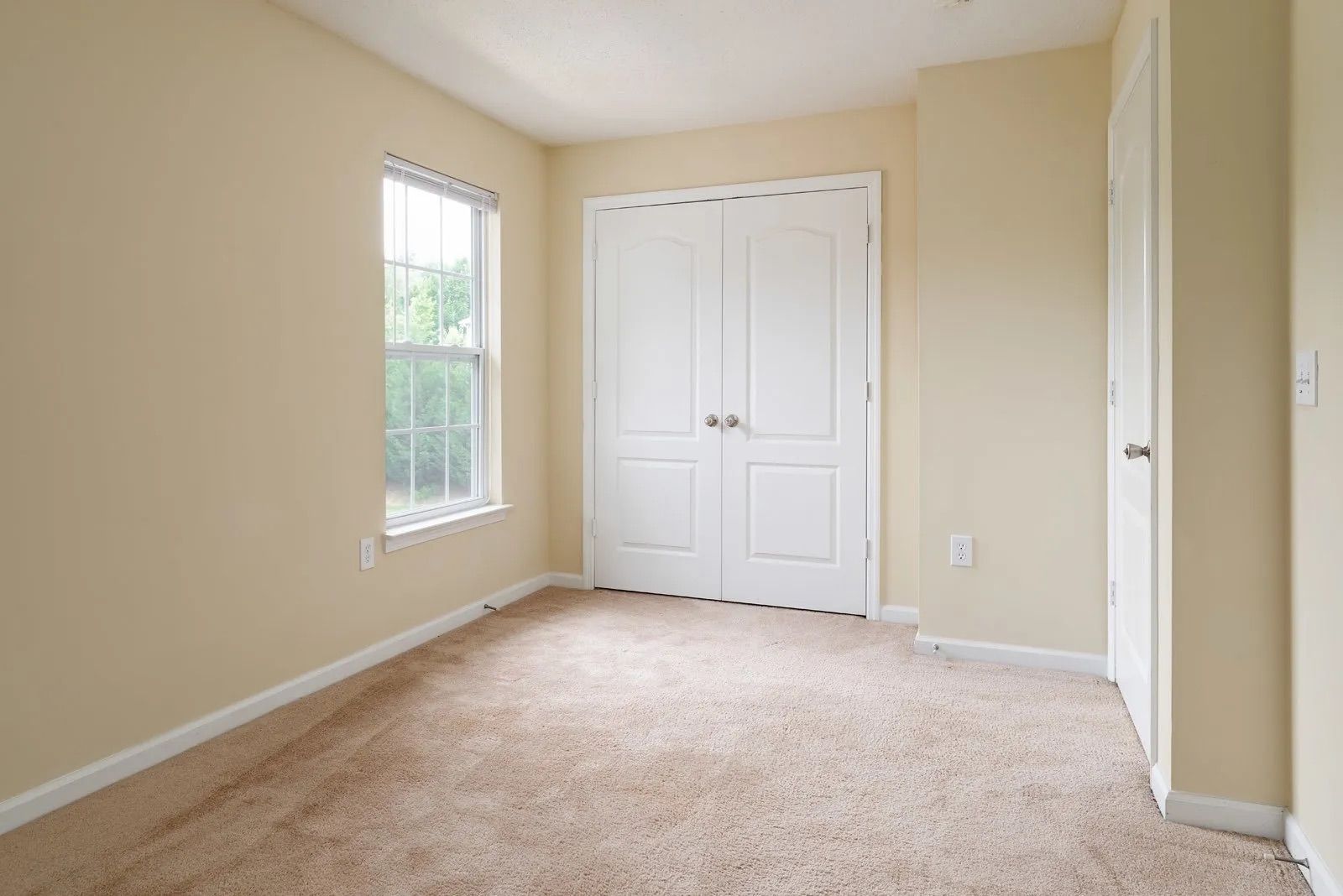 Empty bedroom with tan carpet, beige walls, and white closet doors. Window on the left, door on the right.