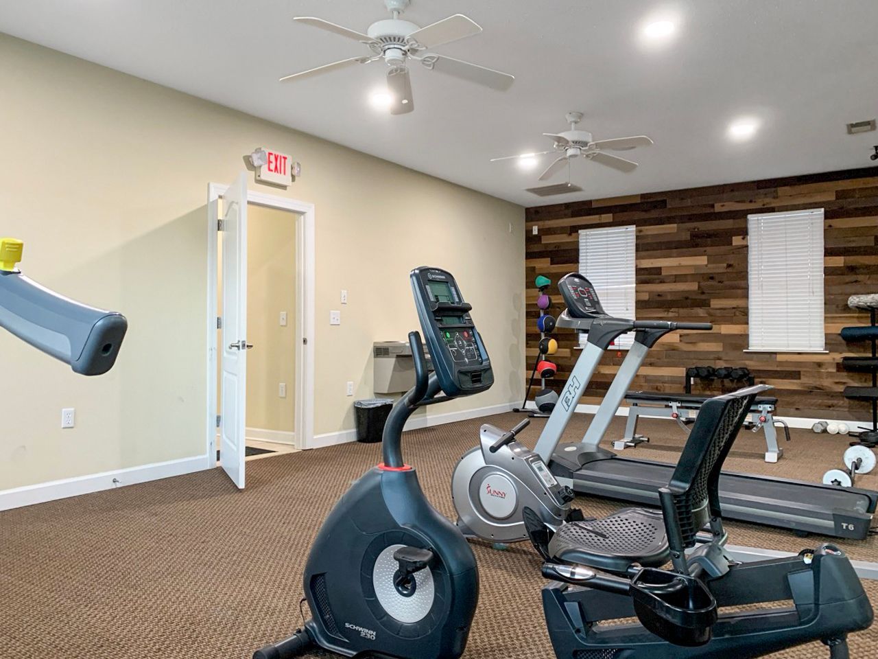 Gym with exercise equipment, including stationary bikes and treadmills. Beige walls, brown carpet.