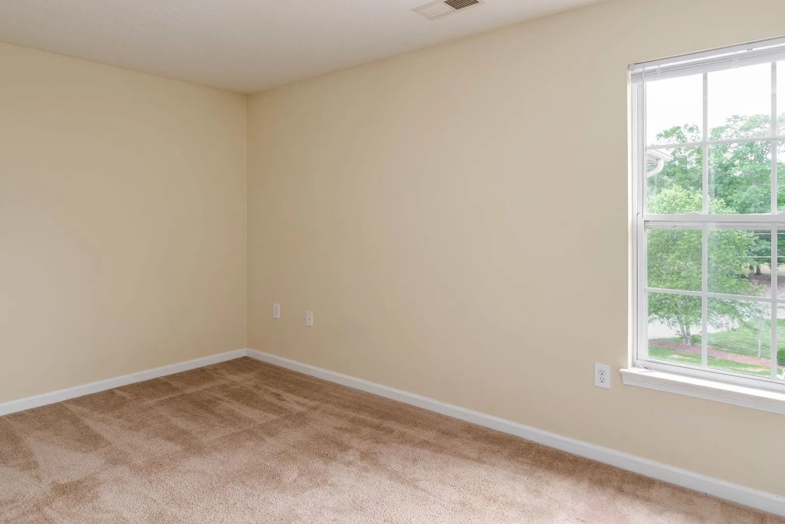 Empty room with beige walls, brown carpet, and a window with a view of greenery.