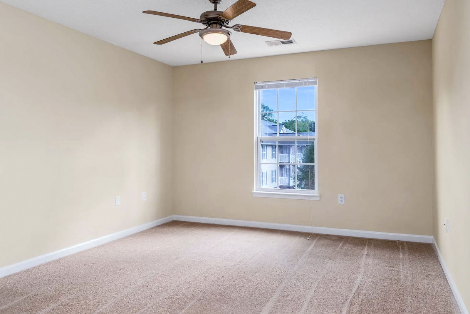 Empty beige-walled room with a window, ceiling fan, and carpeted floor.