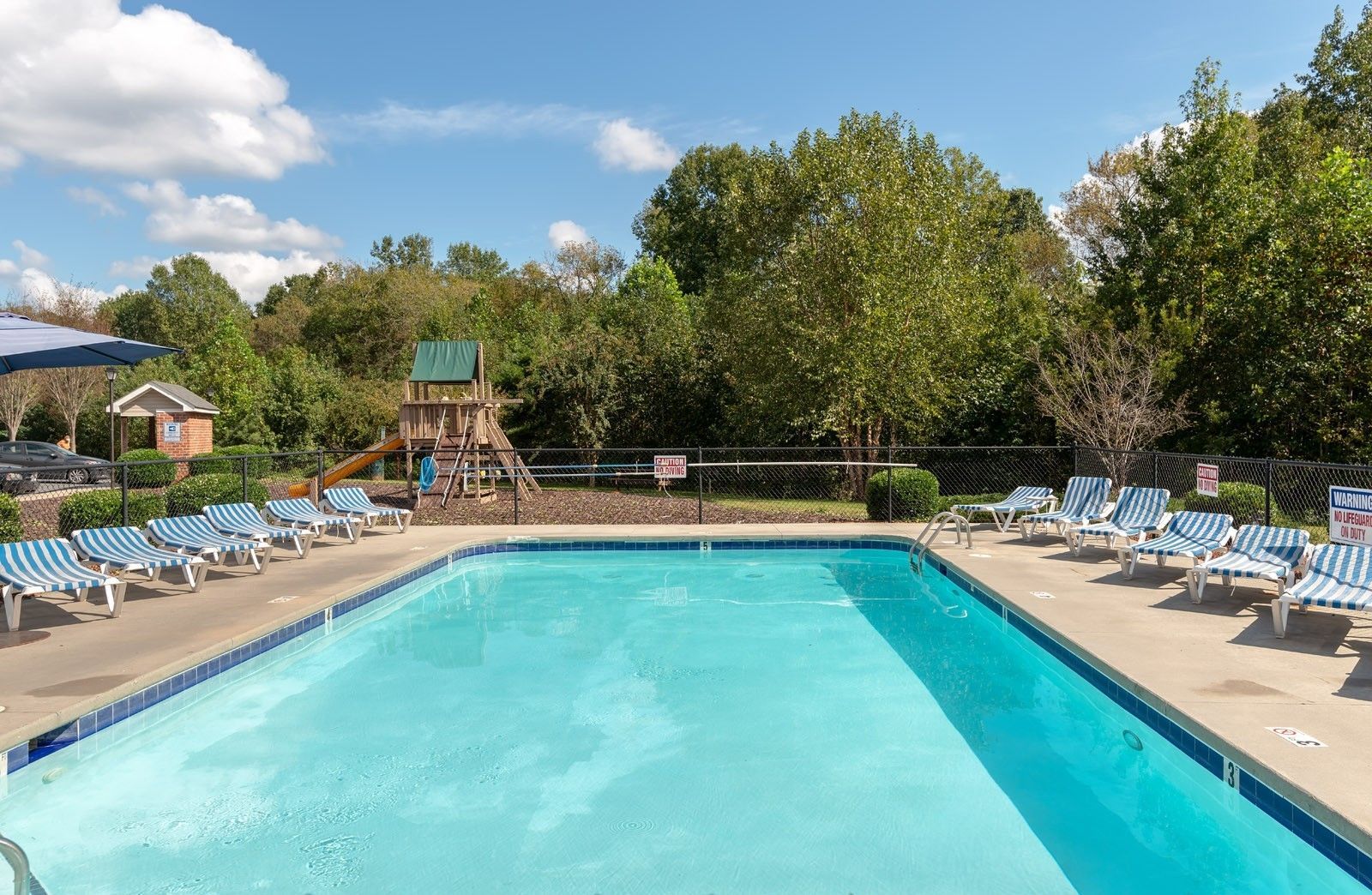 Swimming pool with lounge chairs, trees, and a playground under a blue sky.