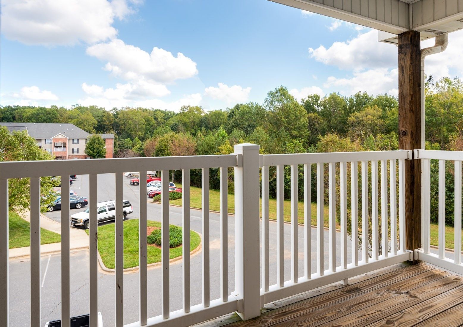 Balcony overlooking a parking lot and trees on a sunny day. White railing, wooden floor.