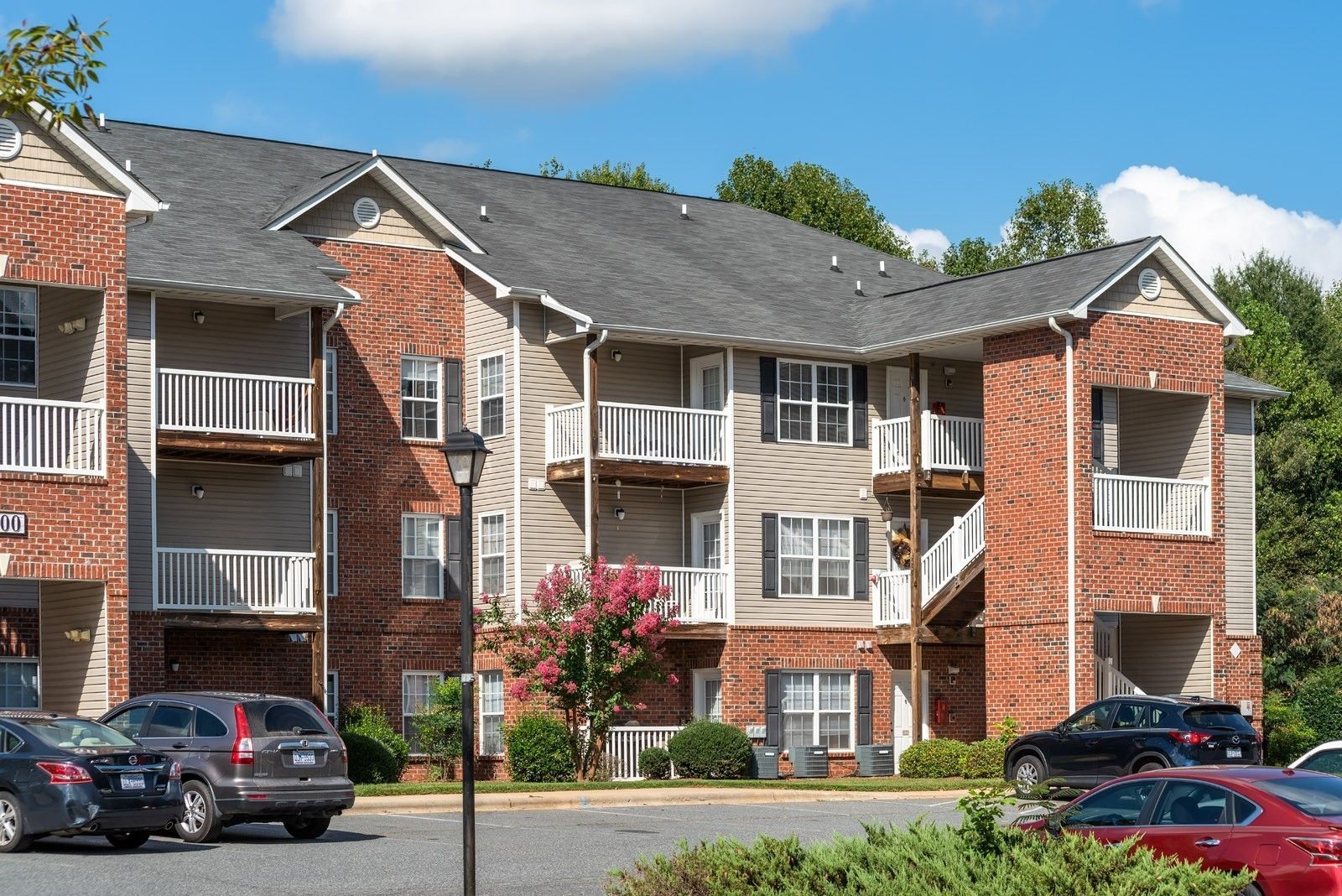 Apartment building with brick facade, balconies, and parked cars on a sunny day.