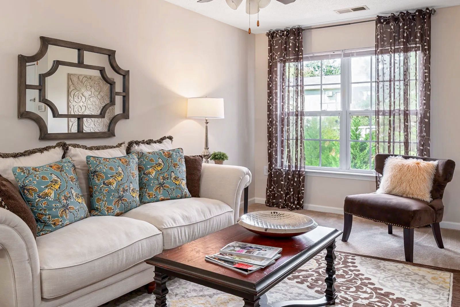 Living room with off-white couch, patterned pillows, coffee table, brown curtains, and decorative mirror.