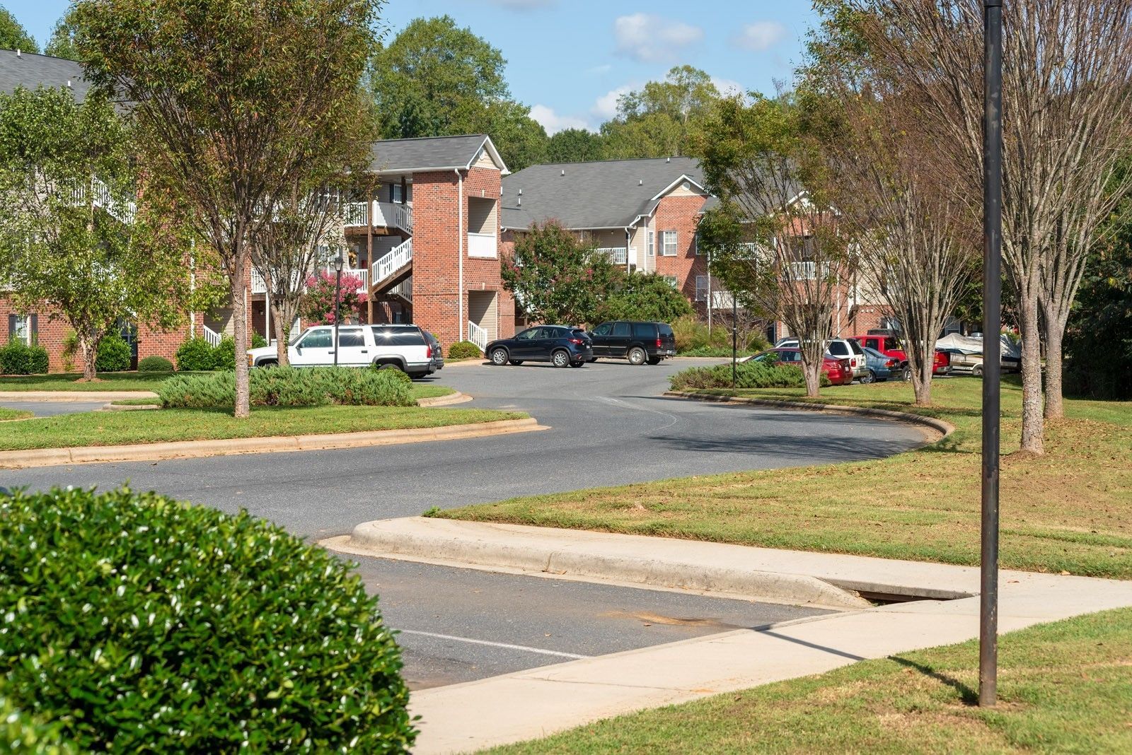 Apartment buildings with brick facade and gray roofs, vehicles parked nearby, greenery in foreground and background.