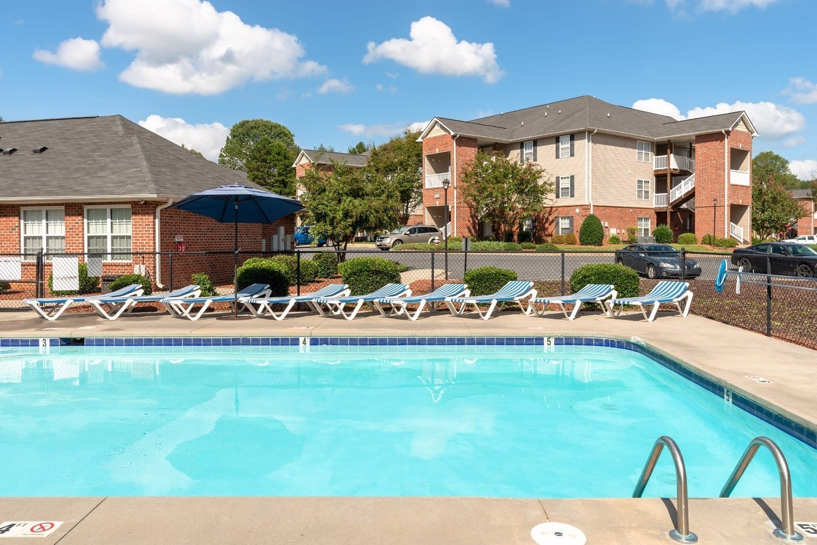 Poolside view of apartment complex with lounge chairs and blue water. Sunny day.