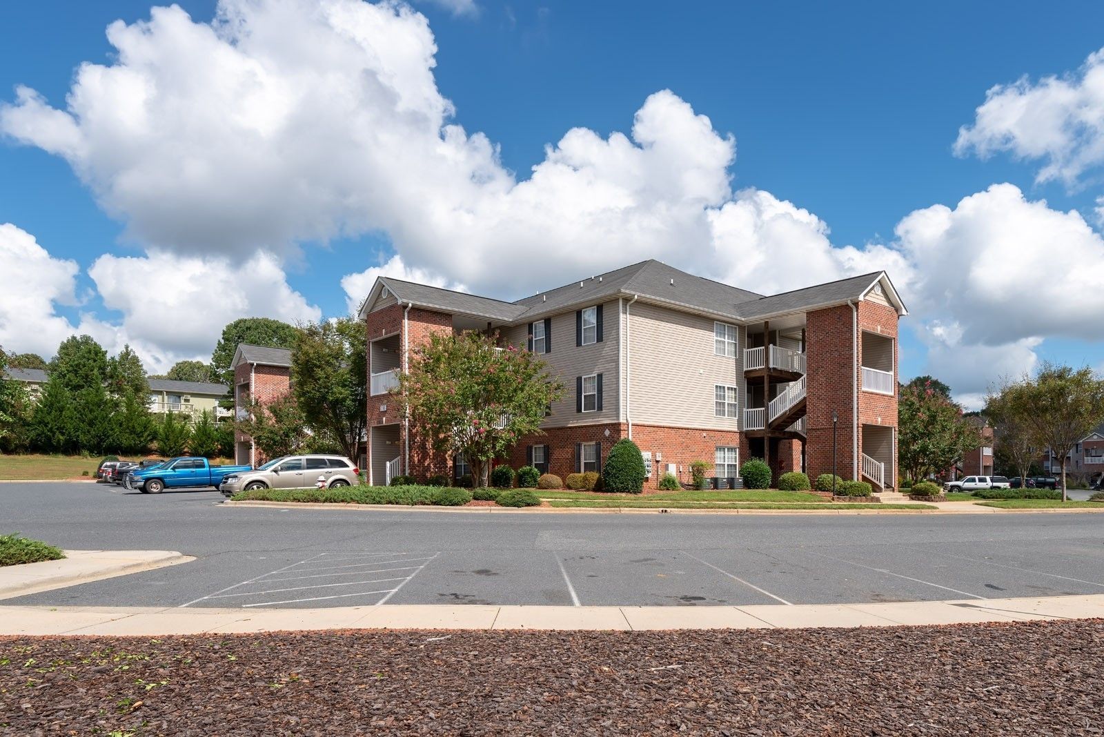 Apartment building with brick accents and parking lot under a blue sky with fluffy clouds.