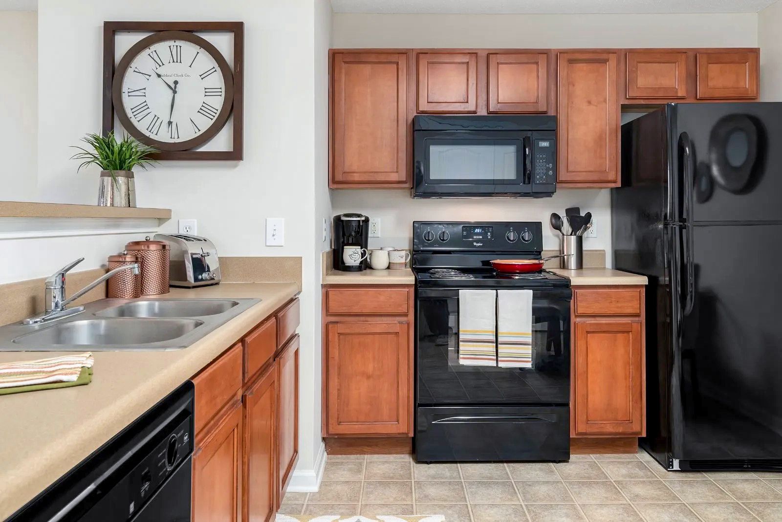 Kitchen with brown cabinets, black appliances, beige countertops, and a large clock on the wall.