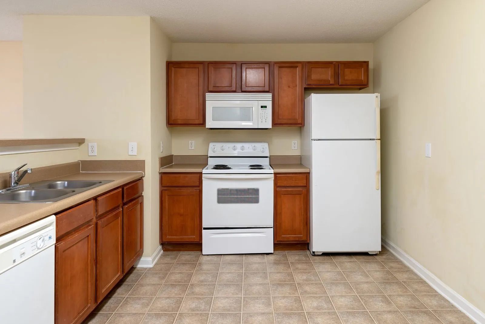 Kitchen with brown cabinets, white appliances (fridge, stove, microwave), and light beige walls and floor.