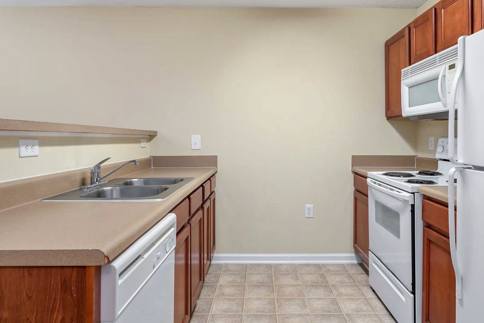 A galley kitchen with brown cabinets, white appliances, and beige countertops and walls.