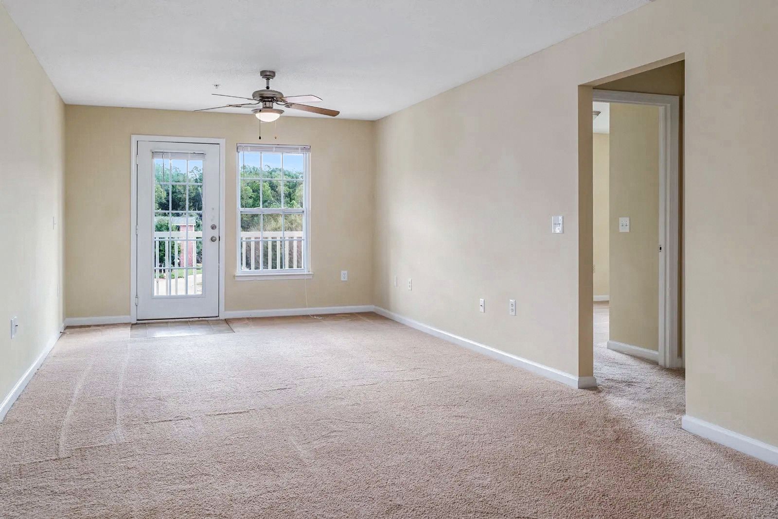 Empty living room with beige walls and carpet, a door to a balcony, and an open doorway to another room.